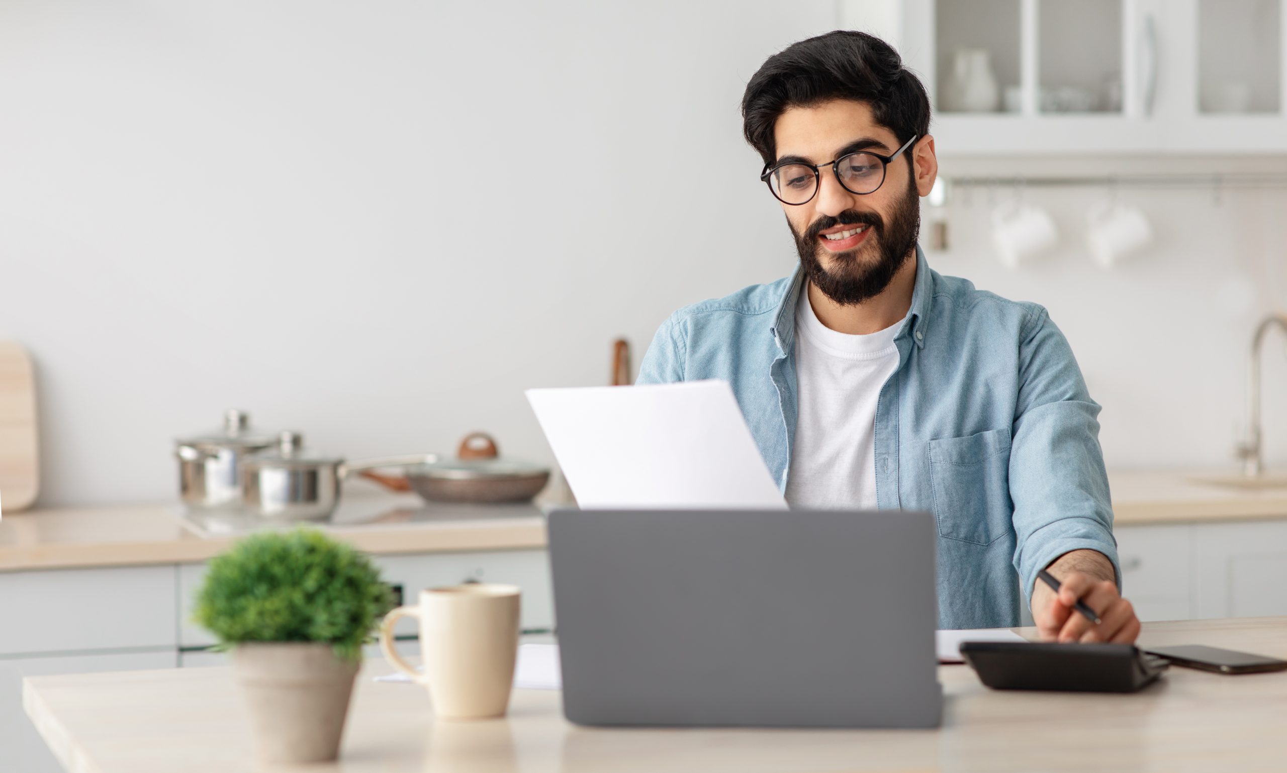 portrait of happy arab guy reading insurance documents at home, sitting at table in kitchen and counting home budget